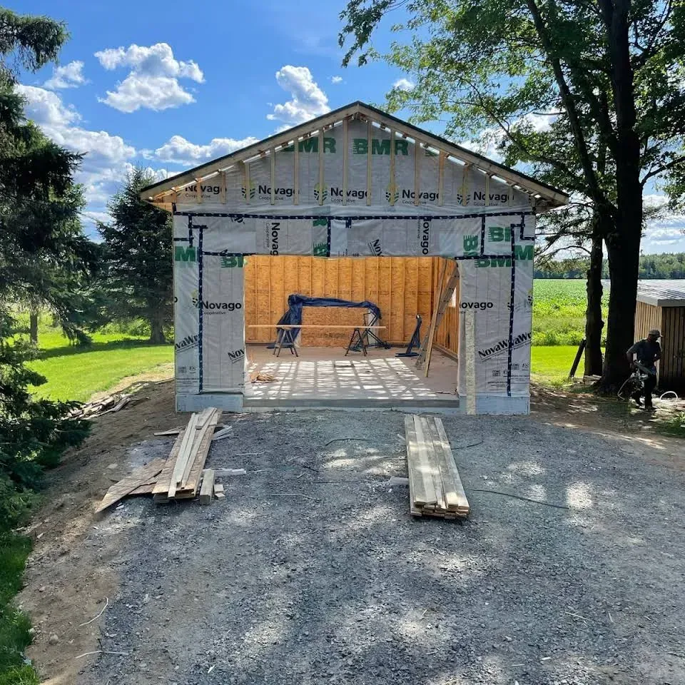 Maison en construction partielle avec charpente apparente et bâche orange à l'intérieur, allée en gravier, cour ensoleillée
