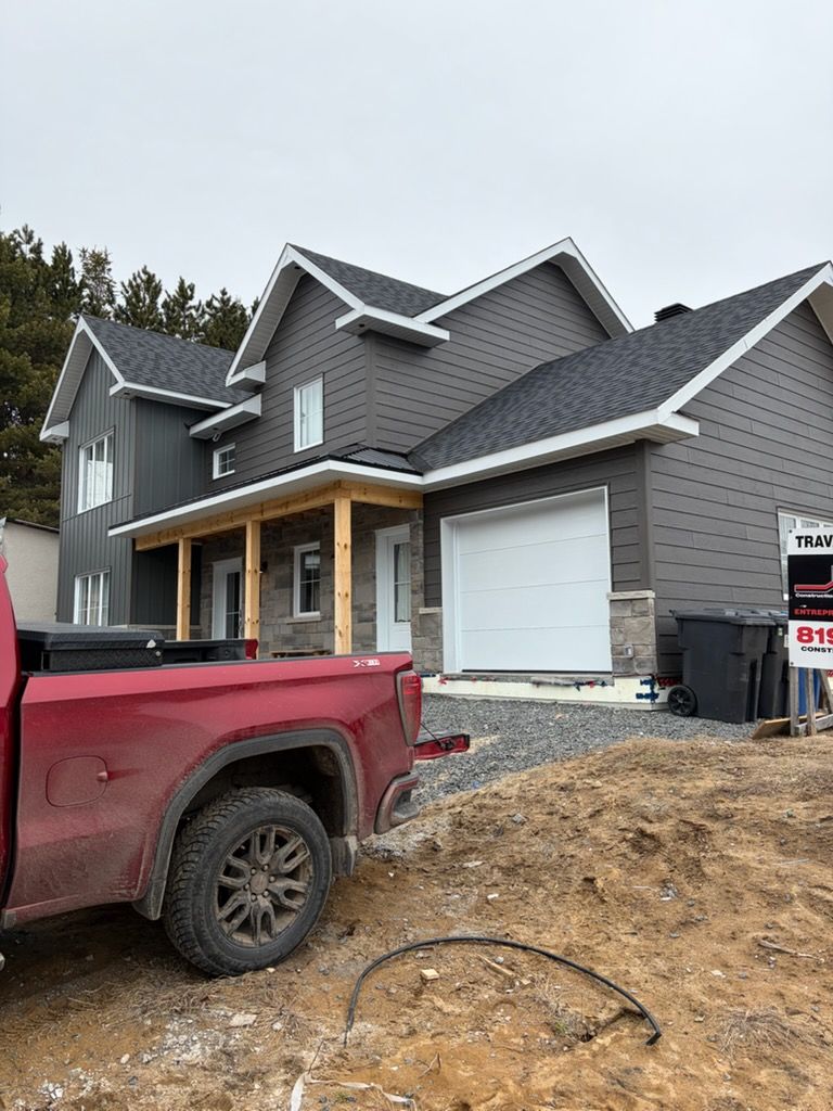 Nouvelle maison grise de banlieue en construction, avec un pick-up rouge et de la terre au premier plan.