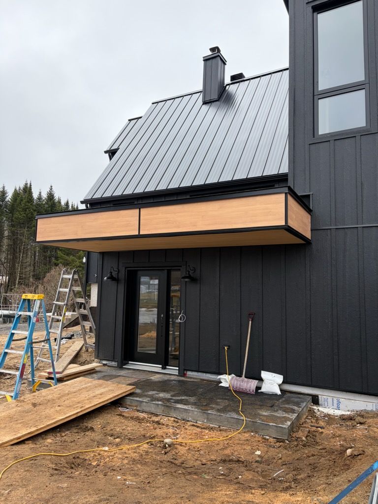 Maison moderne en construction, façade noire, toit en métal gris et balcon en bois.