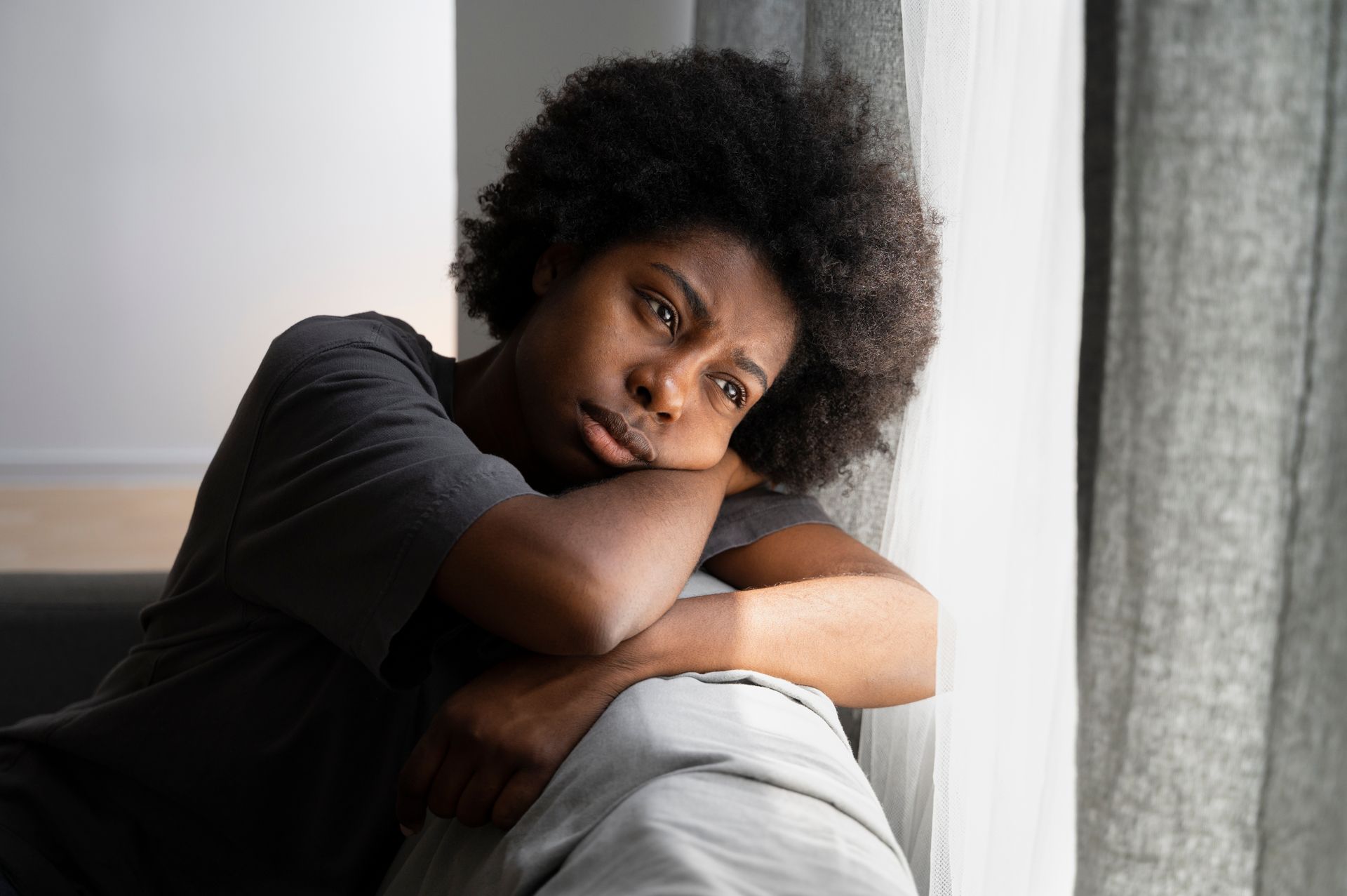 Person resting their head on folded arms by a window, wearing a black shirt and looking pensive.