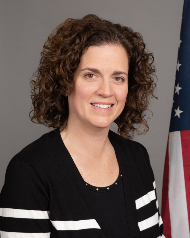 Portrait of a smiling woman in a black top with white stripes, standing before a U.S. flag backdrop