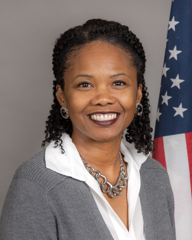 Portrait of a smiling woman in a gray blazer and white blouse, with a U.S. flag in the background