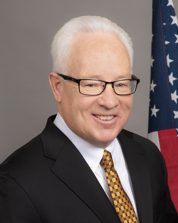 Portrait of a smiling man in a suit and glasses beside a U.S. flag