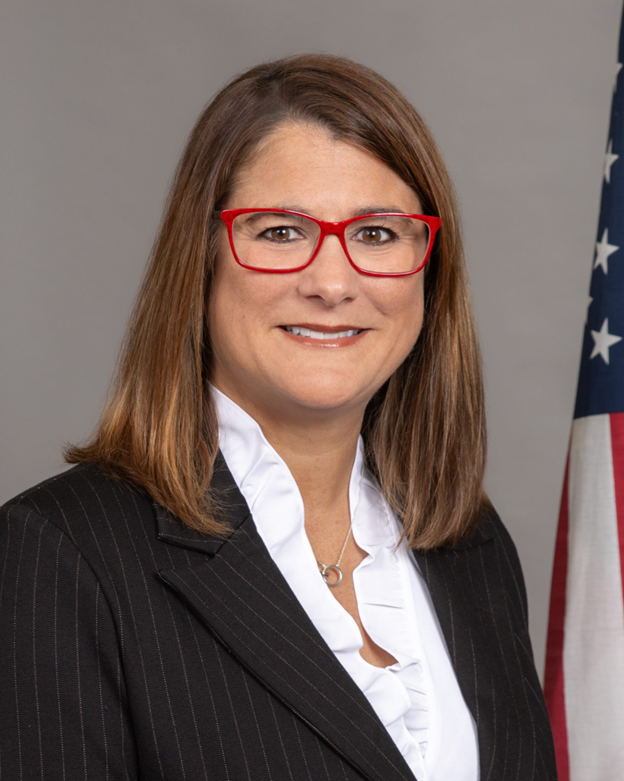 Professional headshot of a woman in a black suit and red glasses, standing before a U.S. flag.