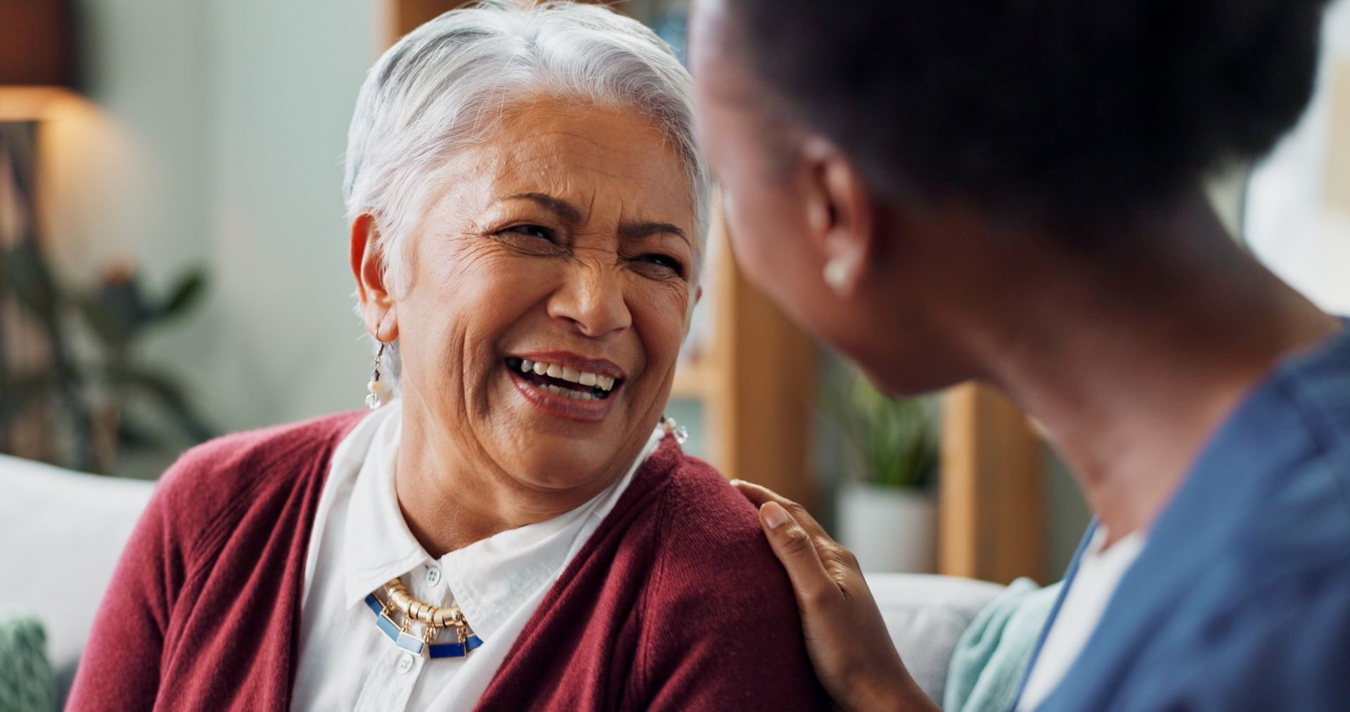 Two people smiling and talking in a cozy living room, one resting a hand on the other’s shoulder.