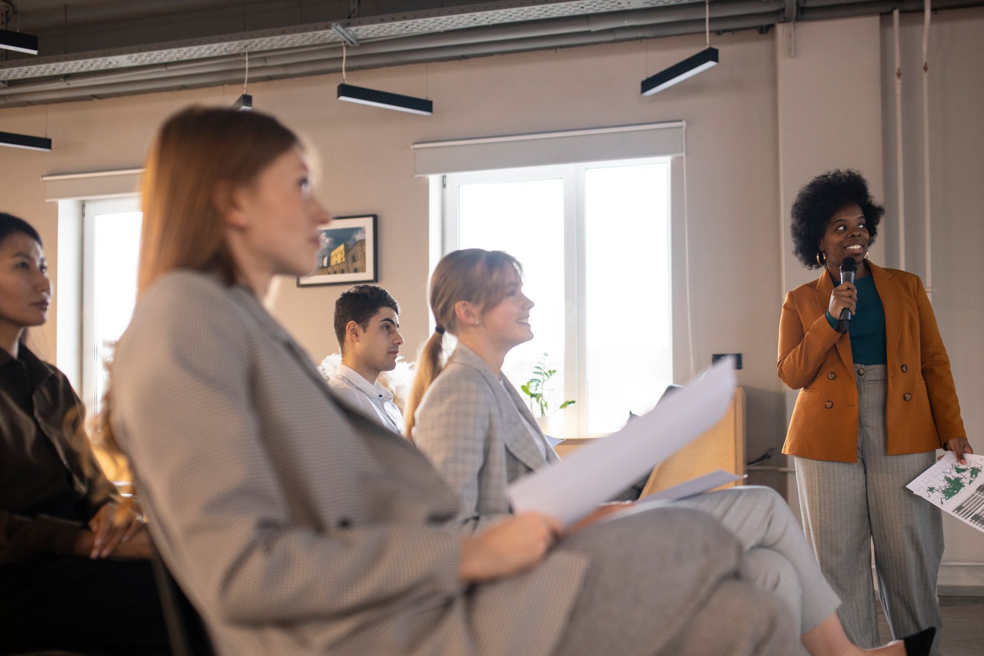 People seated in a bright meeting room listening to a speaker holding a microphone.