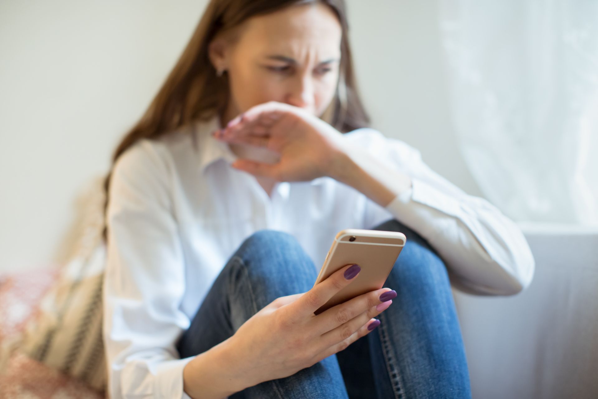 Woman sitting on a couch, covering her mouth while looking at a smartphone, looking distressed