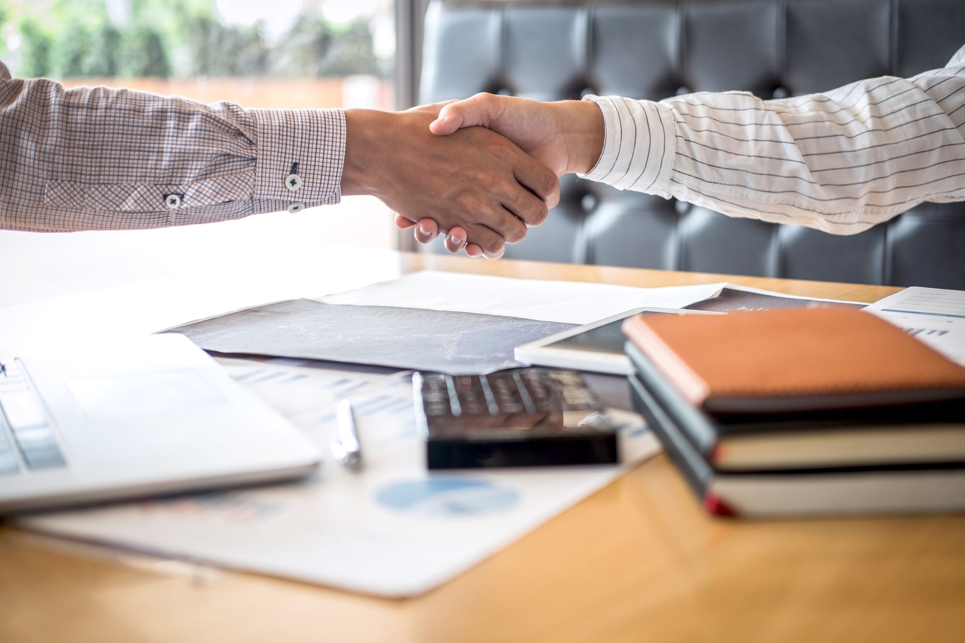 Two people shaking hands across a desk with papers, a laptop, and notebooks in an office setting