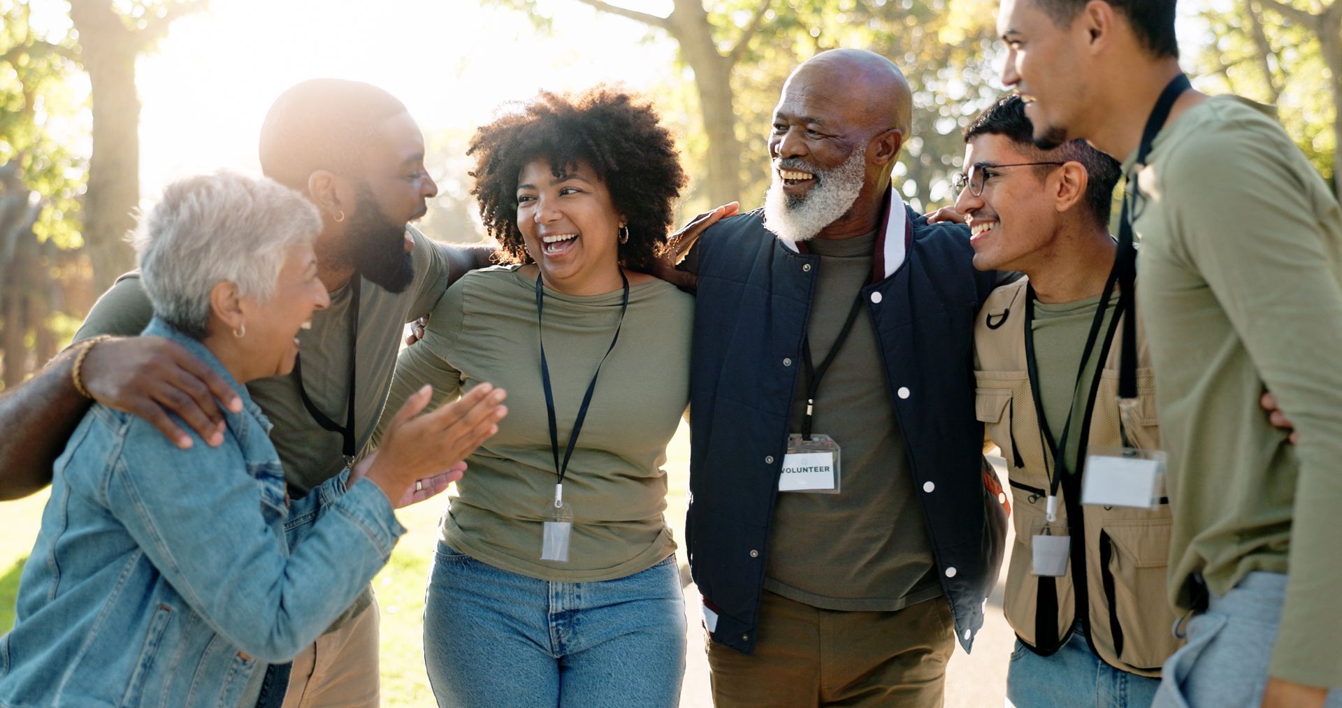 Five people smiling and hugging outdoors in warm sunlight at a park or campus.