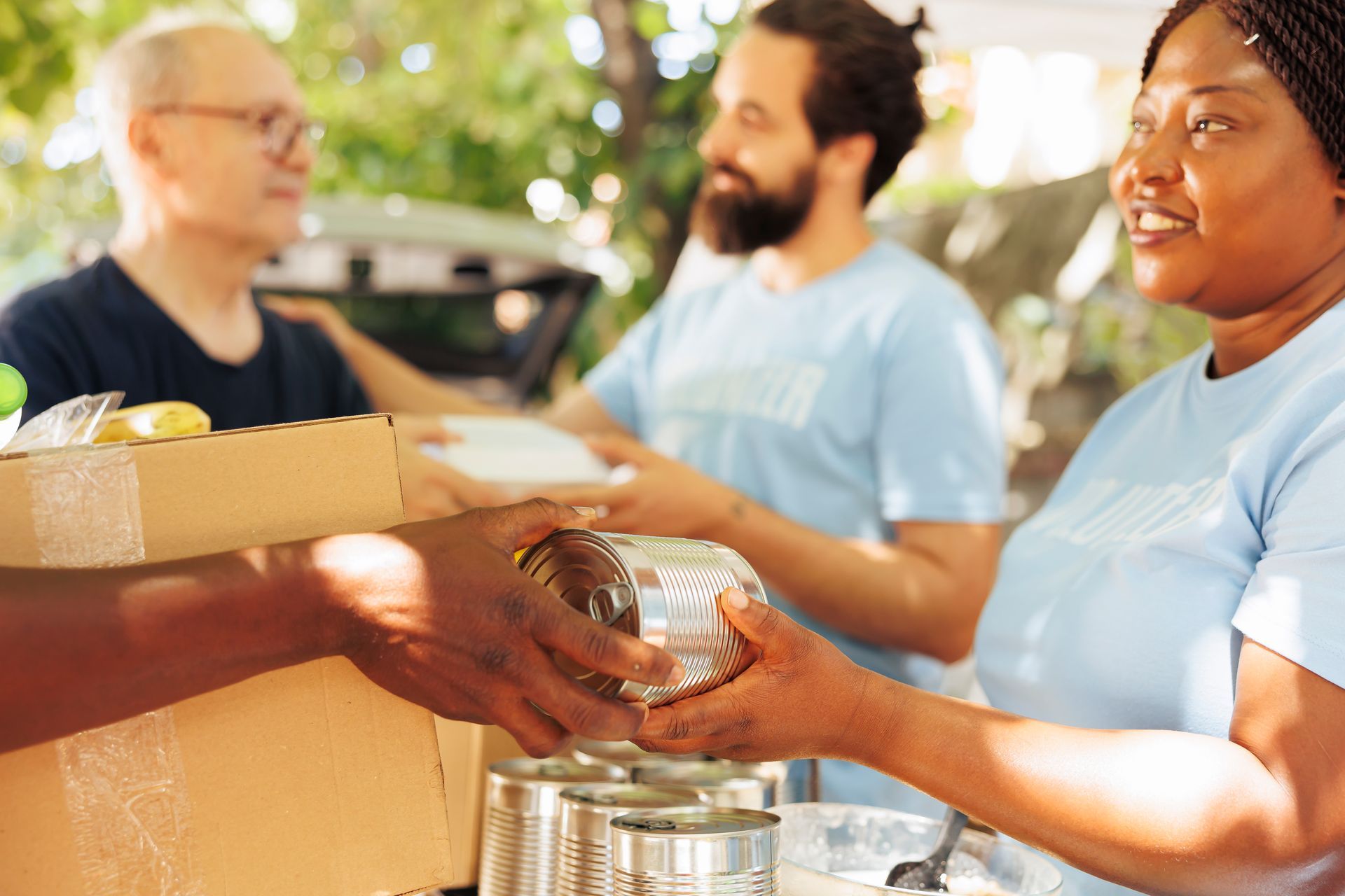 Volunteers passing canned food at an outdoor charity table, with boxes and containers nearby