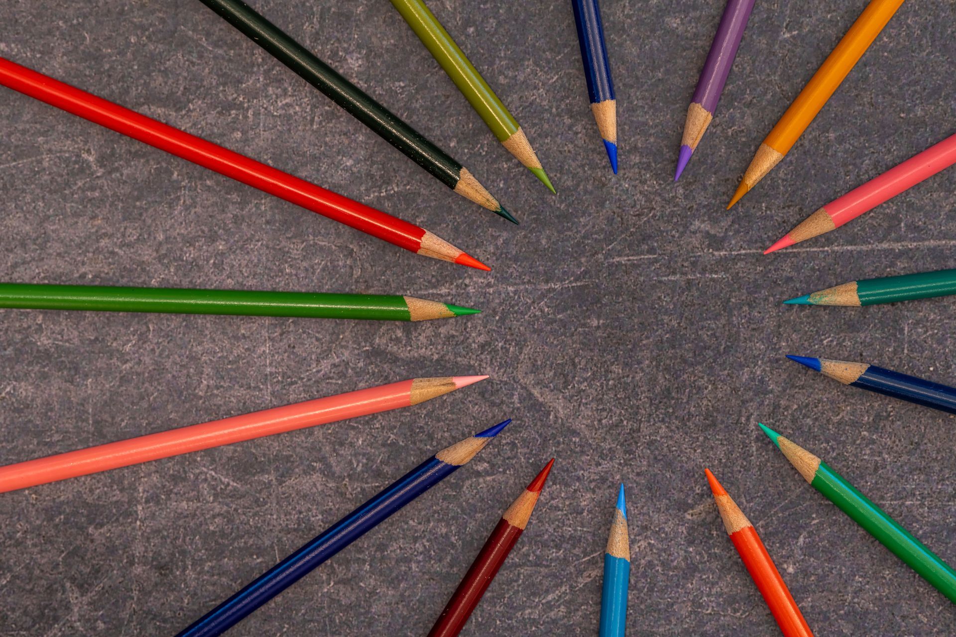 Colored pencils arranged in a circle around an empty center on a textured gray surface