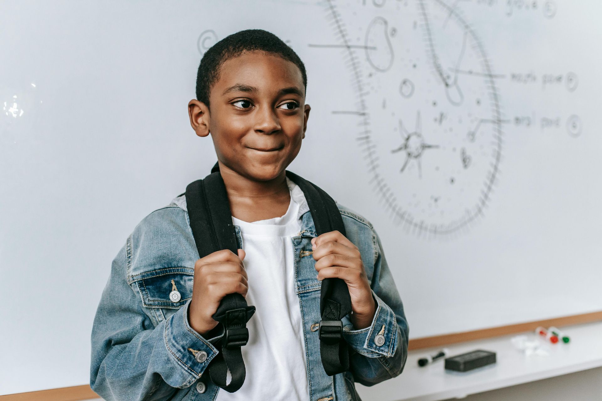 Student with backpack straps smiling in front of a whiteboard in a classroom