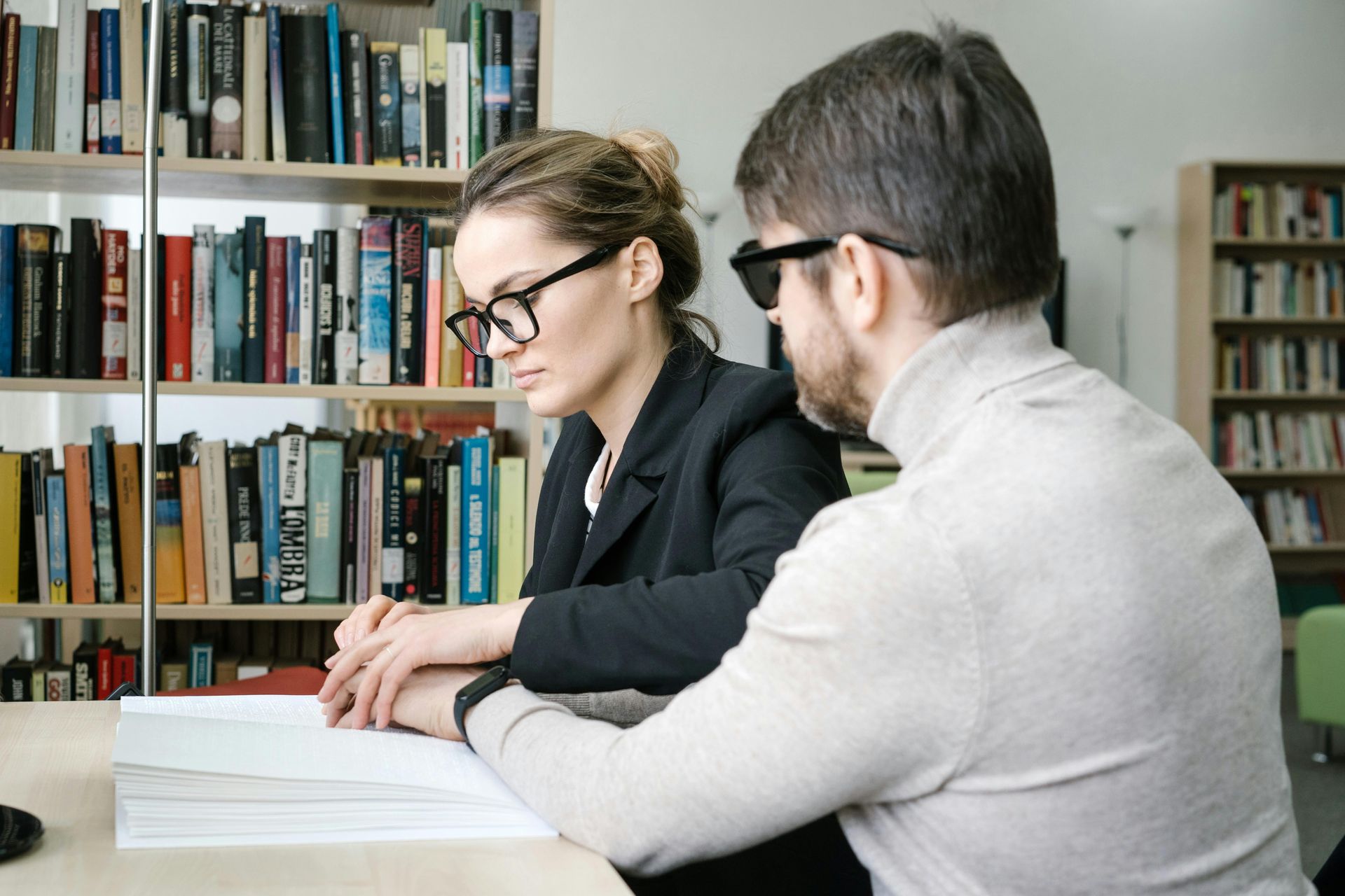 Two people reading at a table in a library, with bookshelves in the background.
