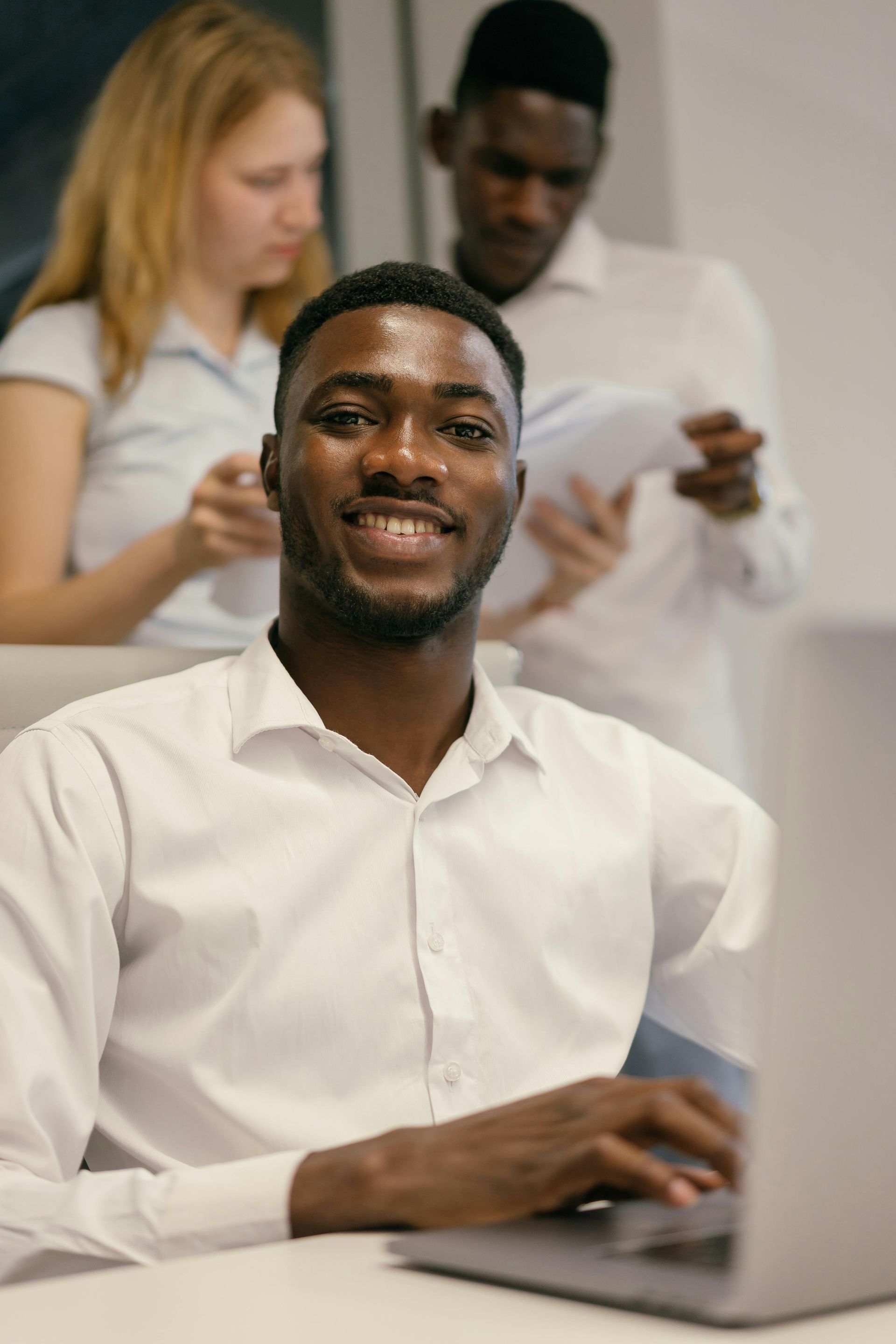 Smiling man in white shirt using a laptop, with two people standing and reading papers behind him