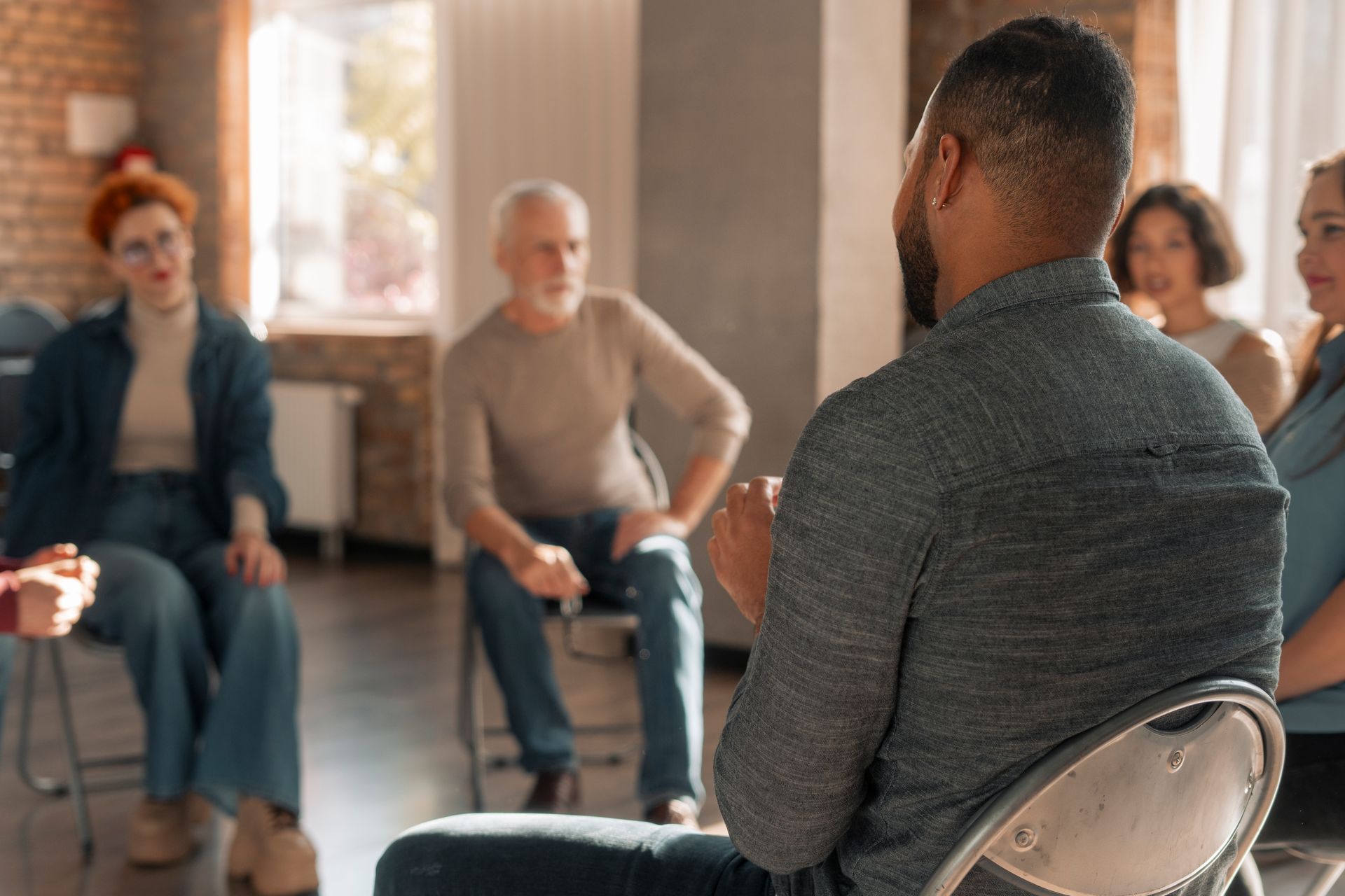 People seated in a small group discussion in a bright room, facing a facilitator.