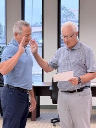 Two men in a room, one gesturing while the other reads from a sheet of paper.