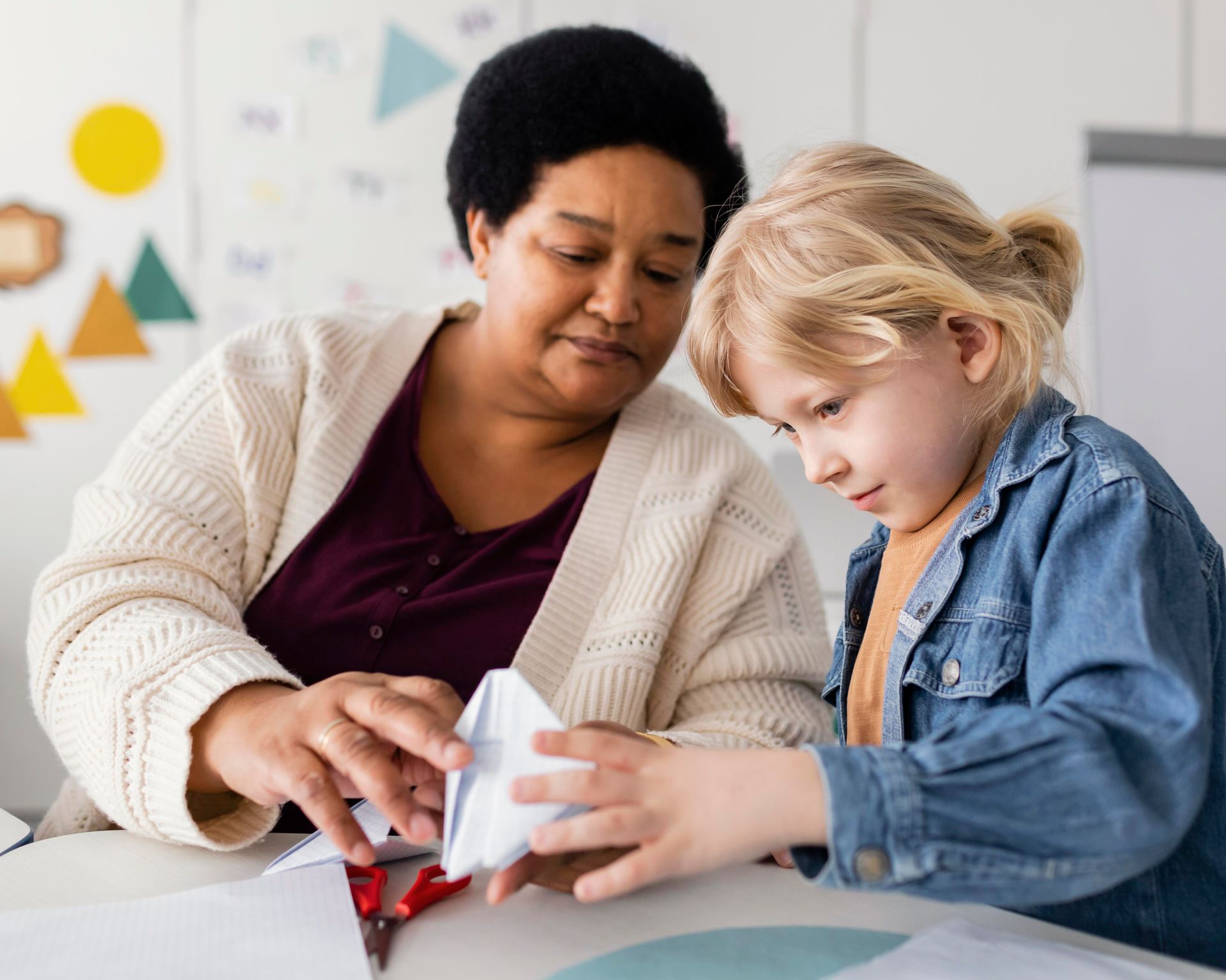 Teacher helping a child build a paper craft at a table in a classroom