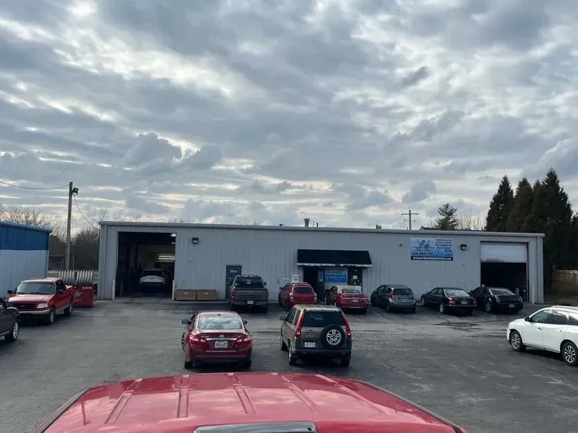 Gray jeep parked outside a white auto repair shop with garage doors and signs on the building