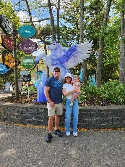 Two people posing by a large purple bird sculpture and colorful signs in a wooded park