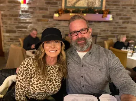 Two people smiling at a restaurant table, with a brick wall and warm lights in the background.