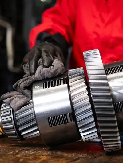 Gloved hands handling shiny metal gears and a shaft on a workbench, with a red jacket in the background