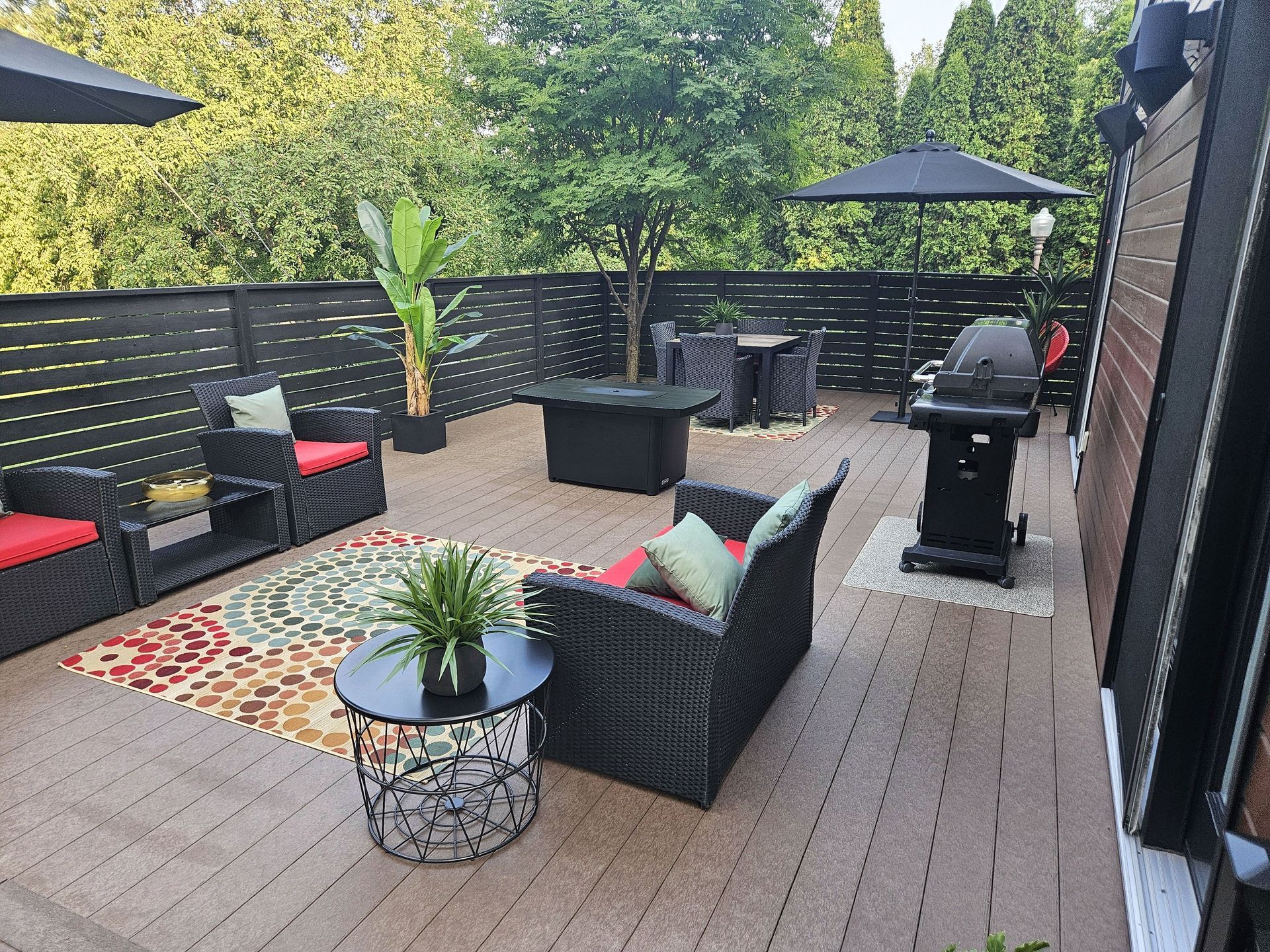 Une terrasse extérieure moderne avec mobilier de jardin en osier, tapis à motifs, foyer, barbecue et plantes en pot.