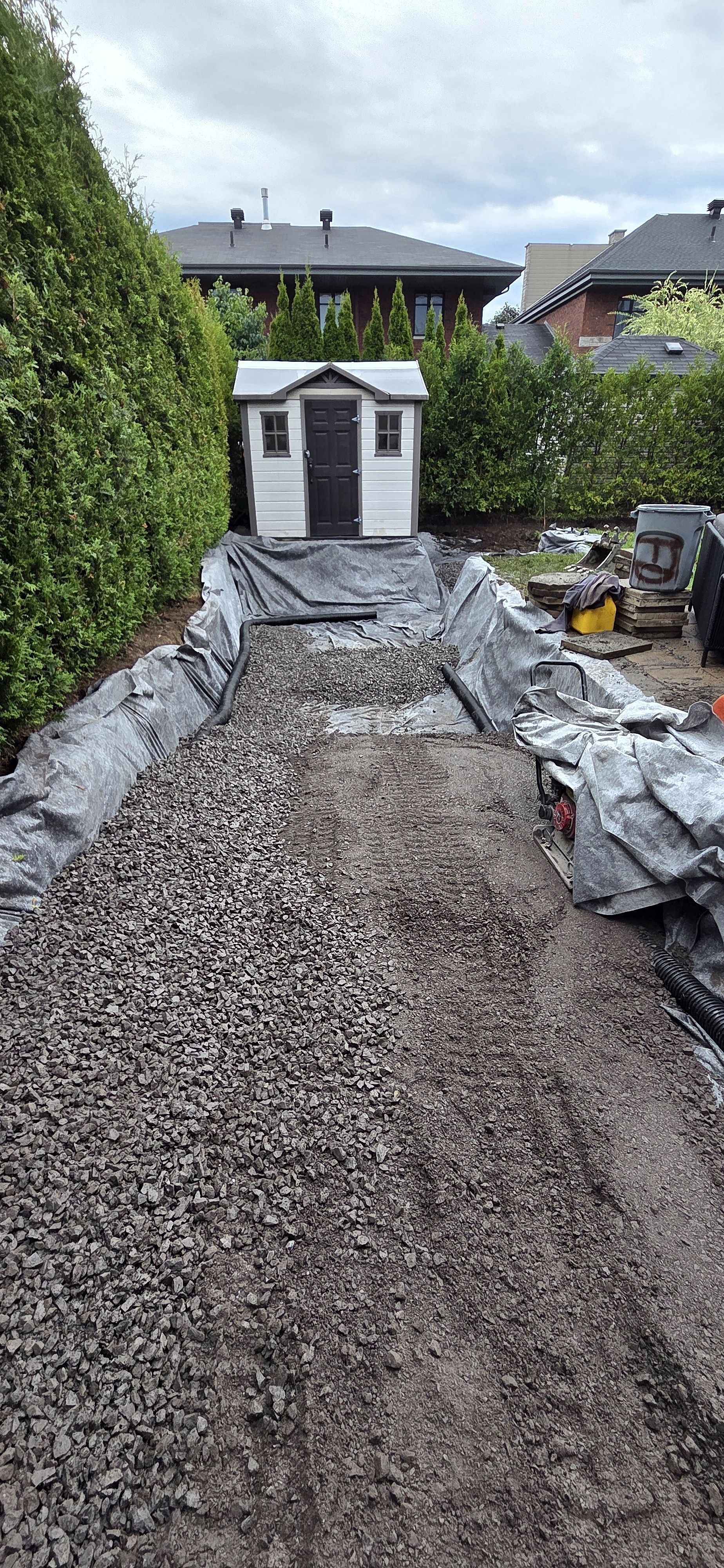Un jardin en travaux avec une tranchée remplie de gravier et tapissée de toile de paillage grise, menant à un petit abri de jardin.