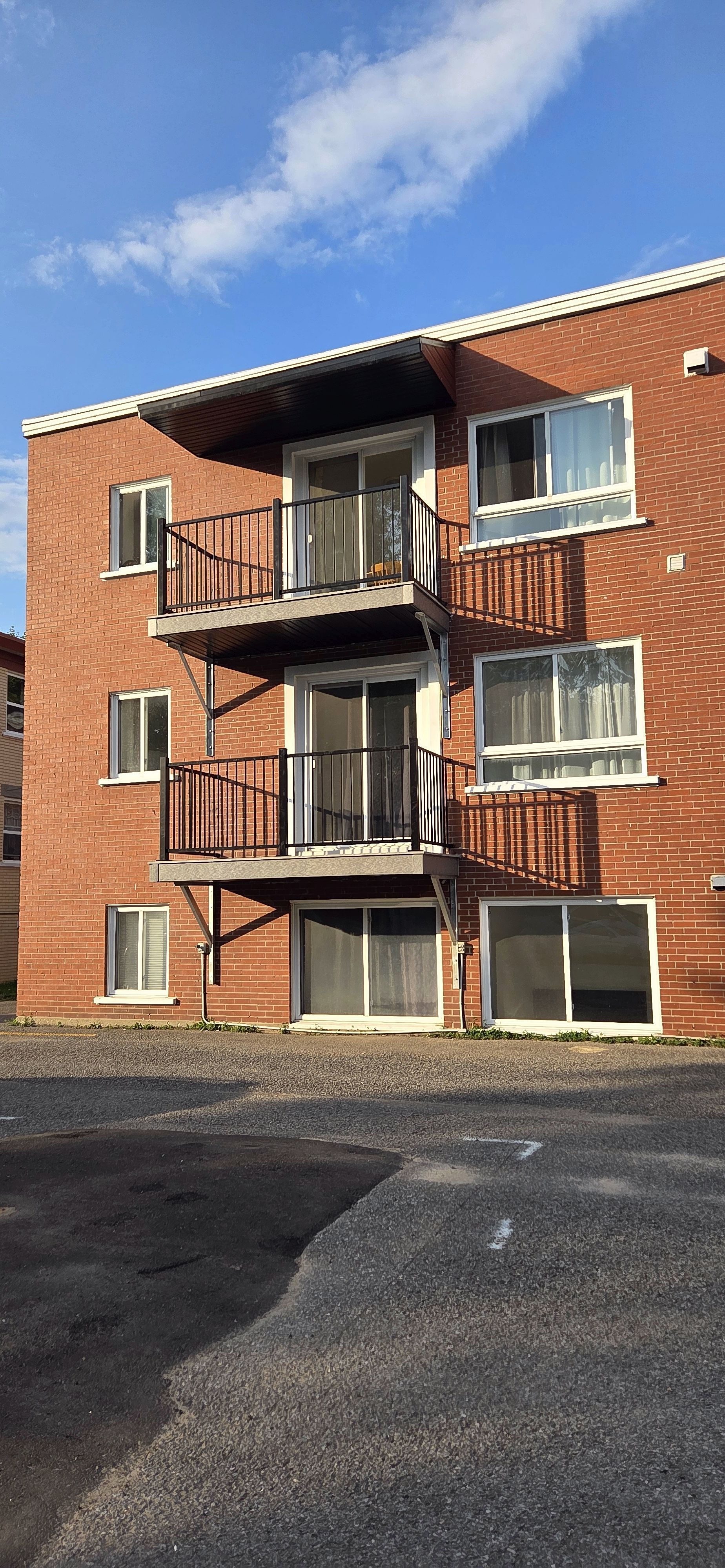 Un immeuble d'appartements en briques de trois étages, avec deux balcons superposés et des portes coulissantes en verre, sous un ciel d'un bleu éclatant.