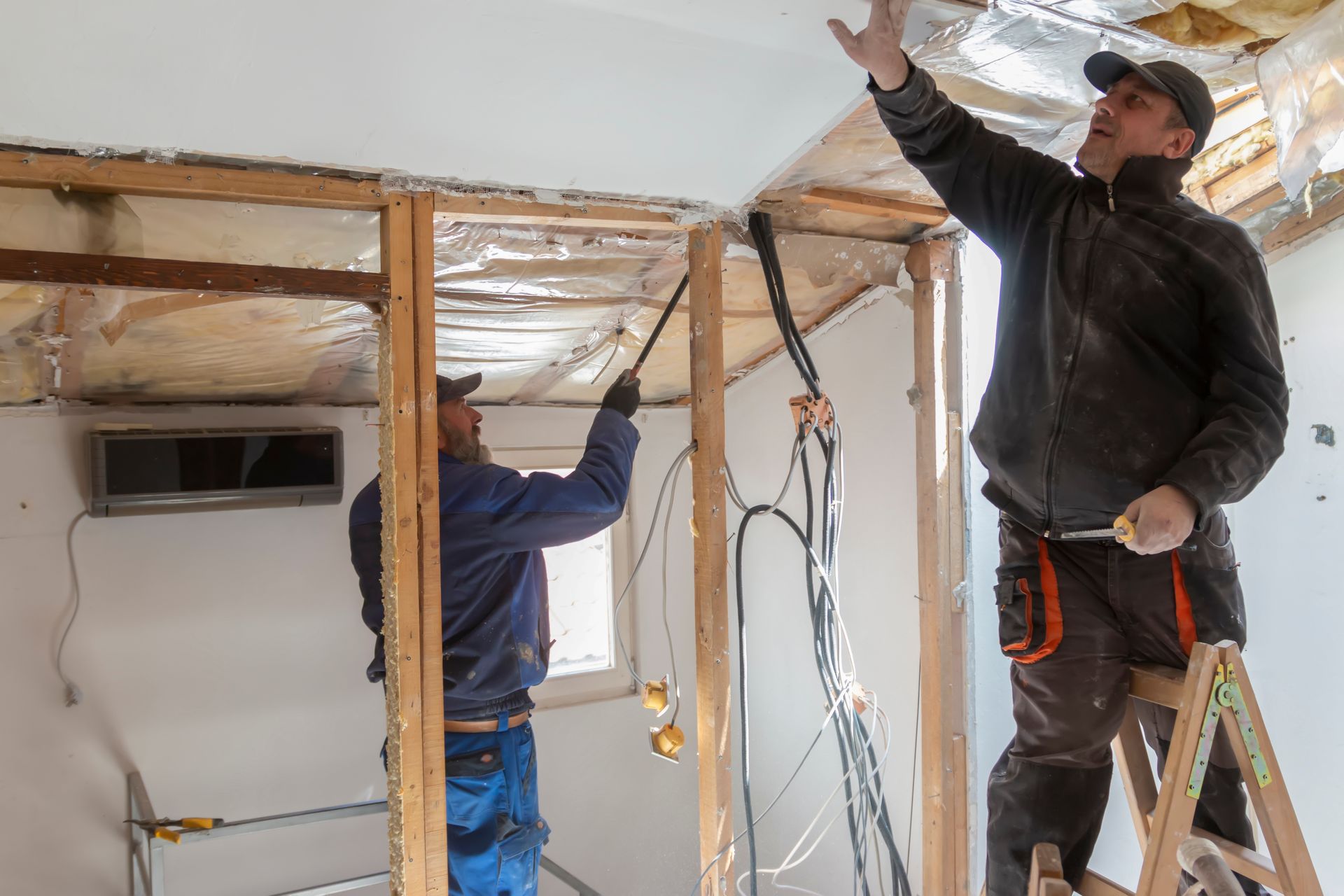 Deux ouvriers du bâtiment en tenue de travail installent des câbles à l'intérieur d'une structure en bois lors de la rénovation d'une pièce.