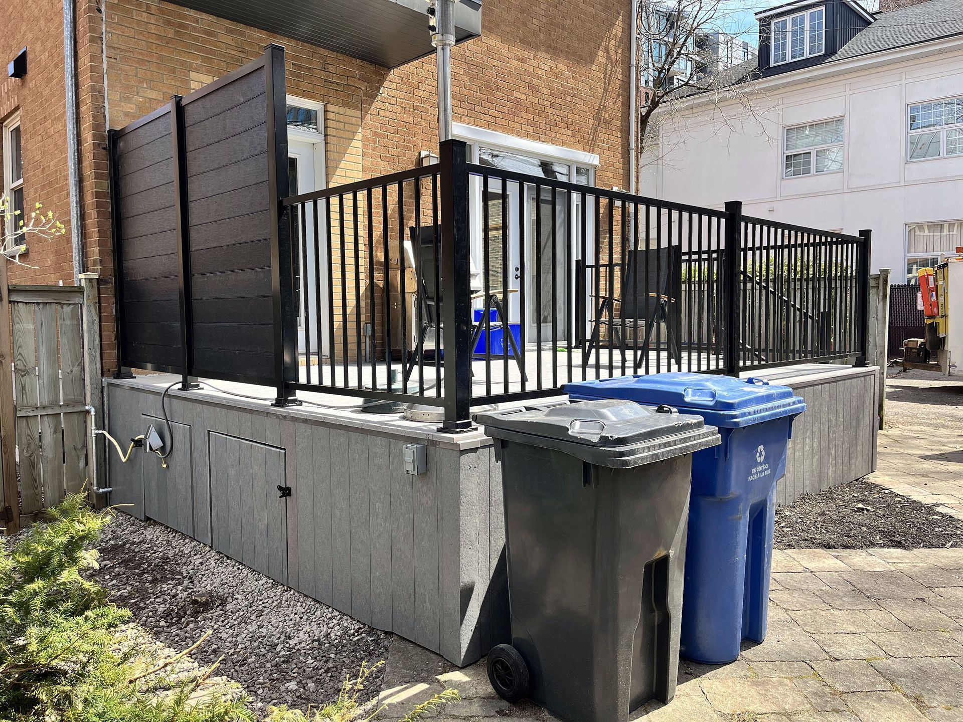 Une terrasse surélevée avec des rambardes en métal noir et des panneaux d'intimité, attenante à un bâtiment en briques, avec deux poubelles devant.