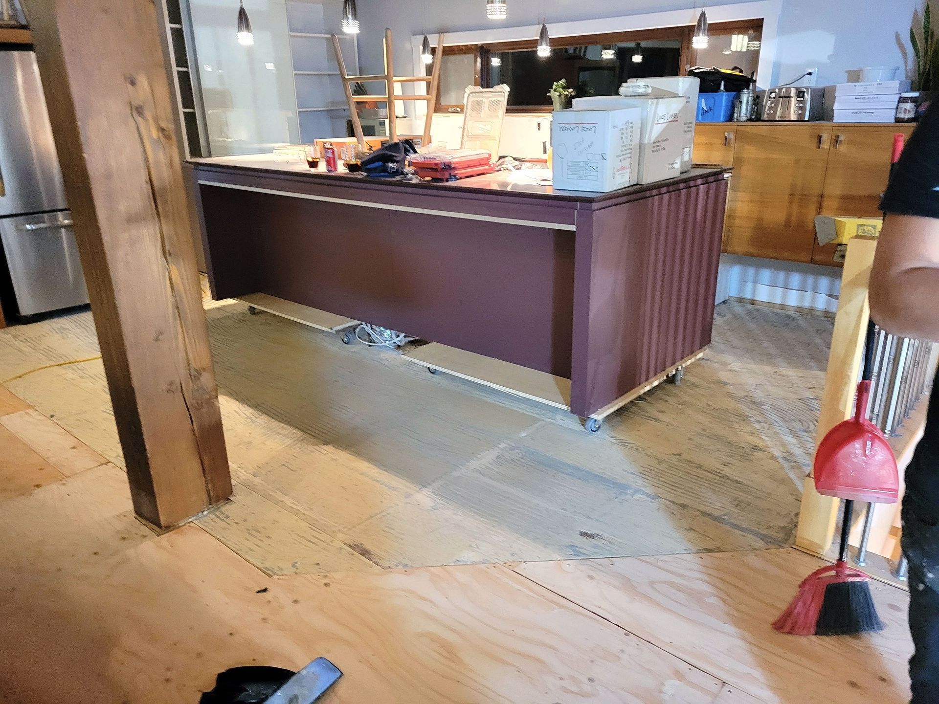 Dans une pièce en rénovation, un îlot de cuisine brun foncé repose sur un sous-plancher en contreplaqué, avec des boîtes blanches posées dessus.