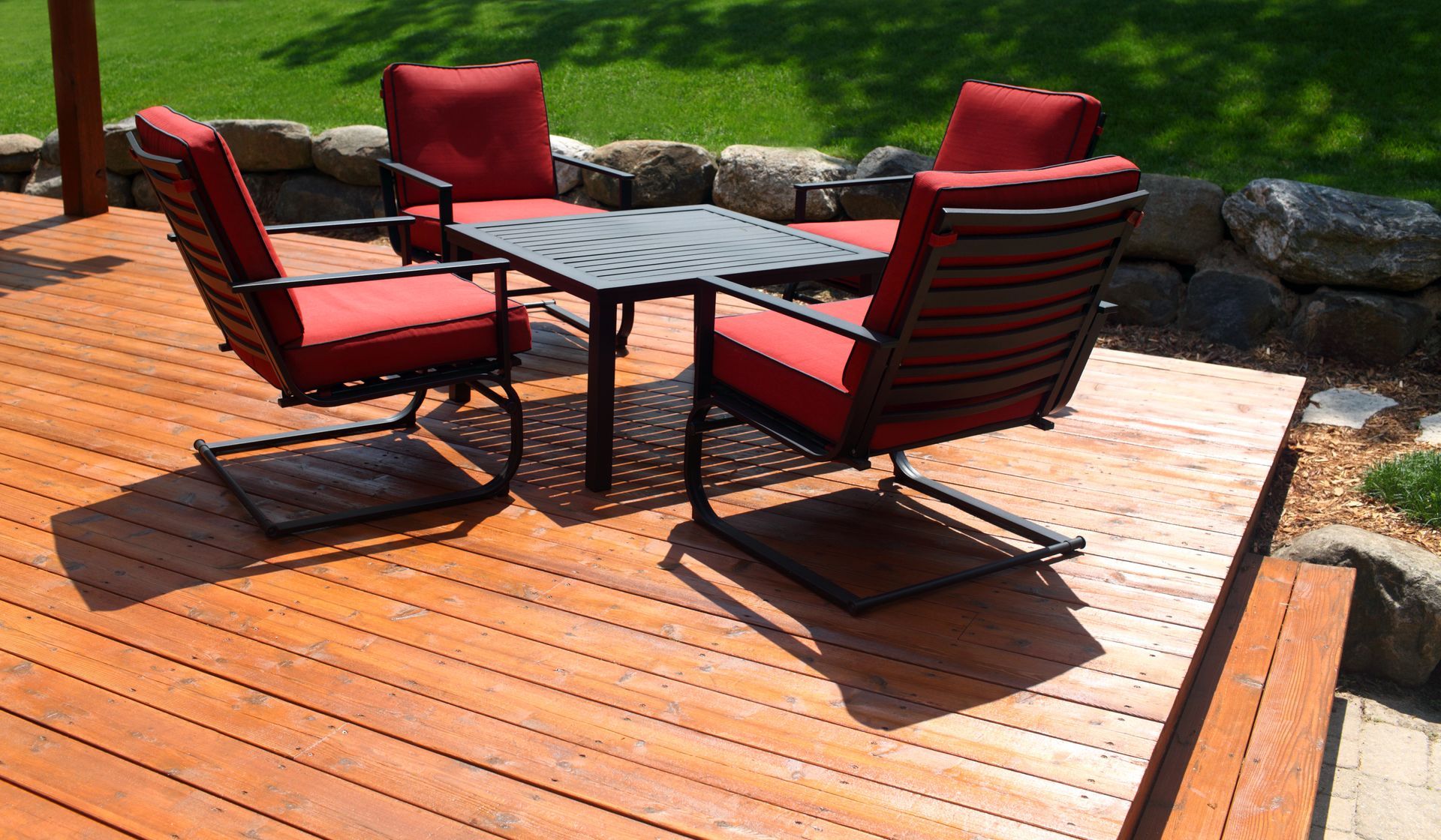 Quatre chaises aux coussins rouges sont disposées autour d'une petite table carrée sur une terrasse en bois dans un jardin.