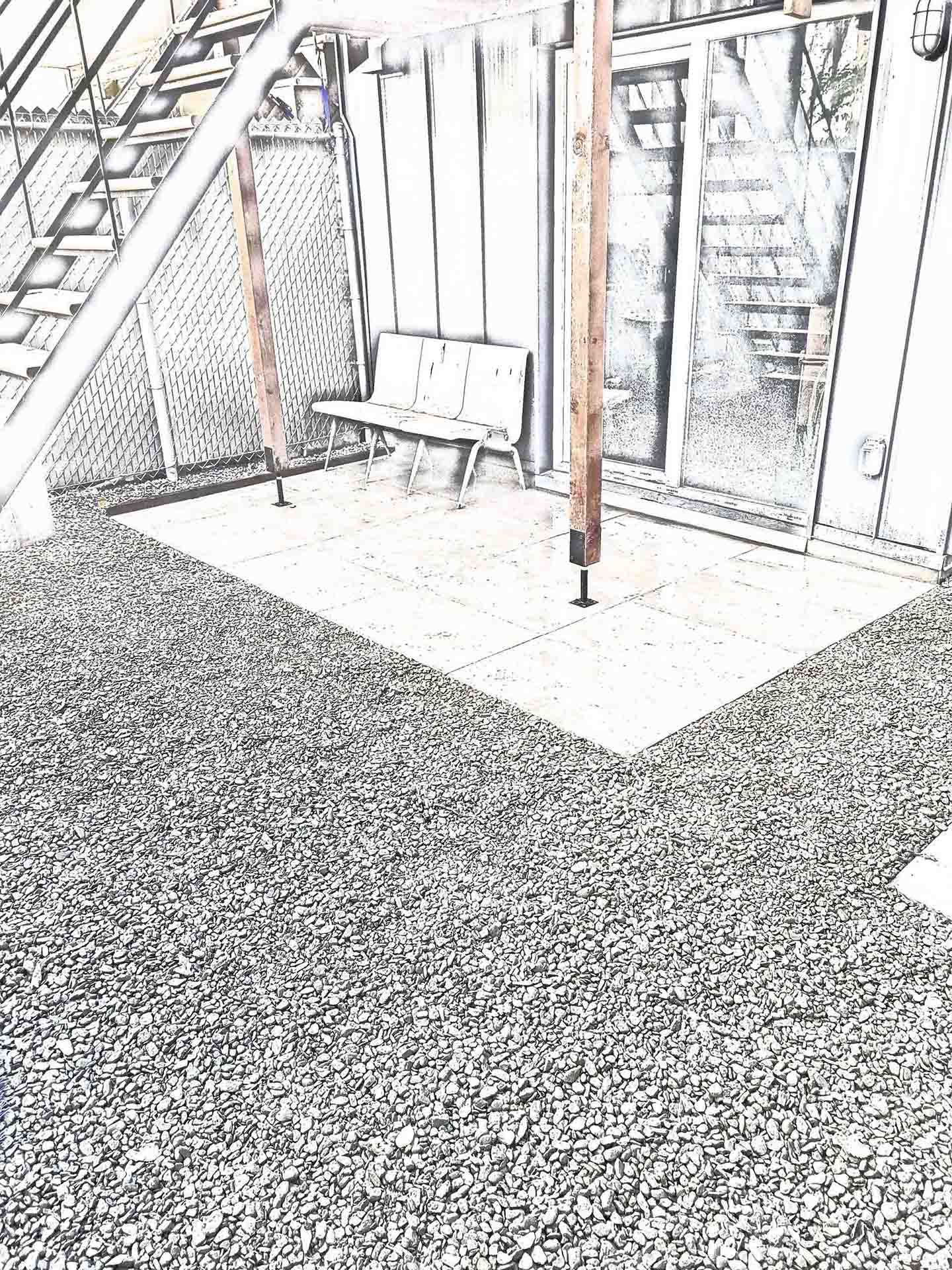 Un patio aux tons gris avec un petit banc sous une structure en bois, adossé à un bâtiment avec un escalier et une porte vitrée.