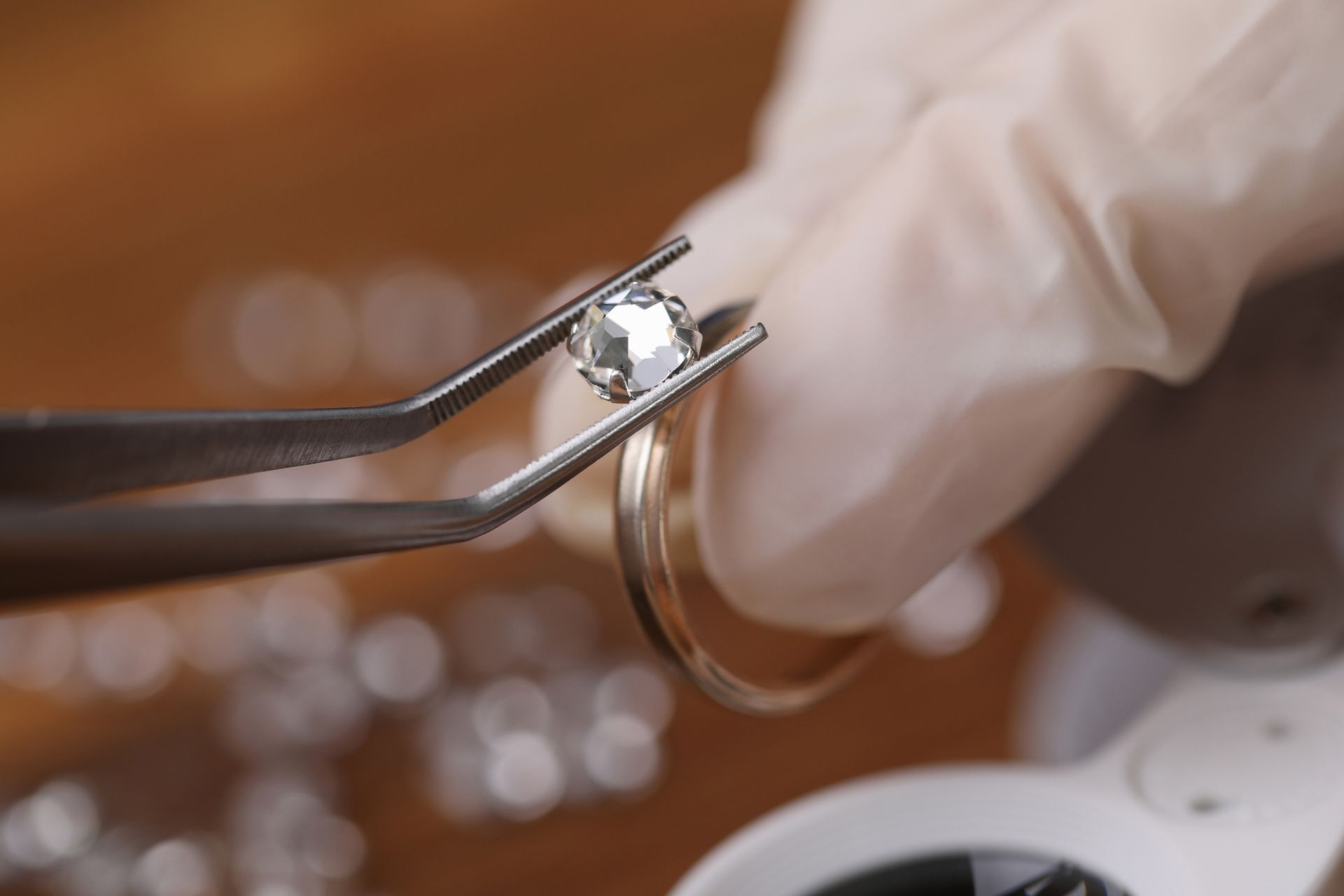 Gloved hand holding a diamond with tweezers above a ring; close-up shot on a wooden surface.