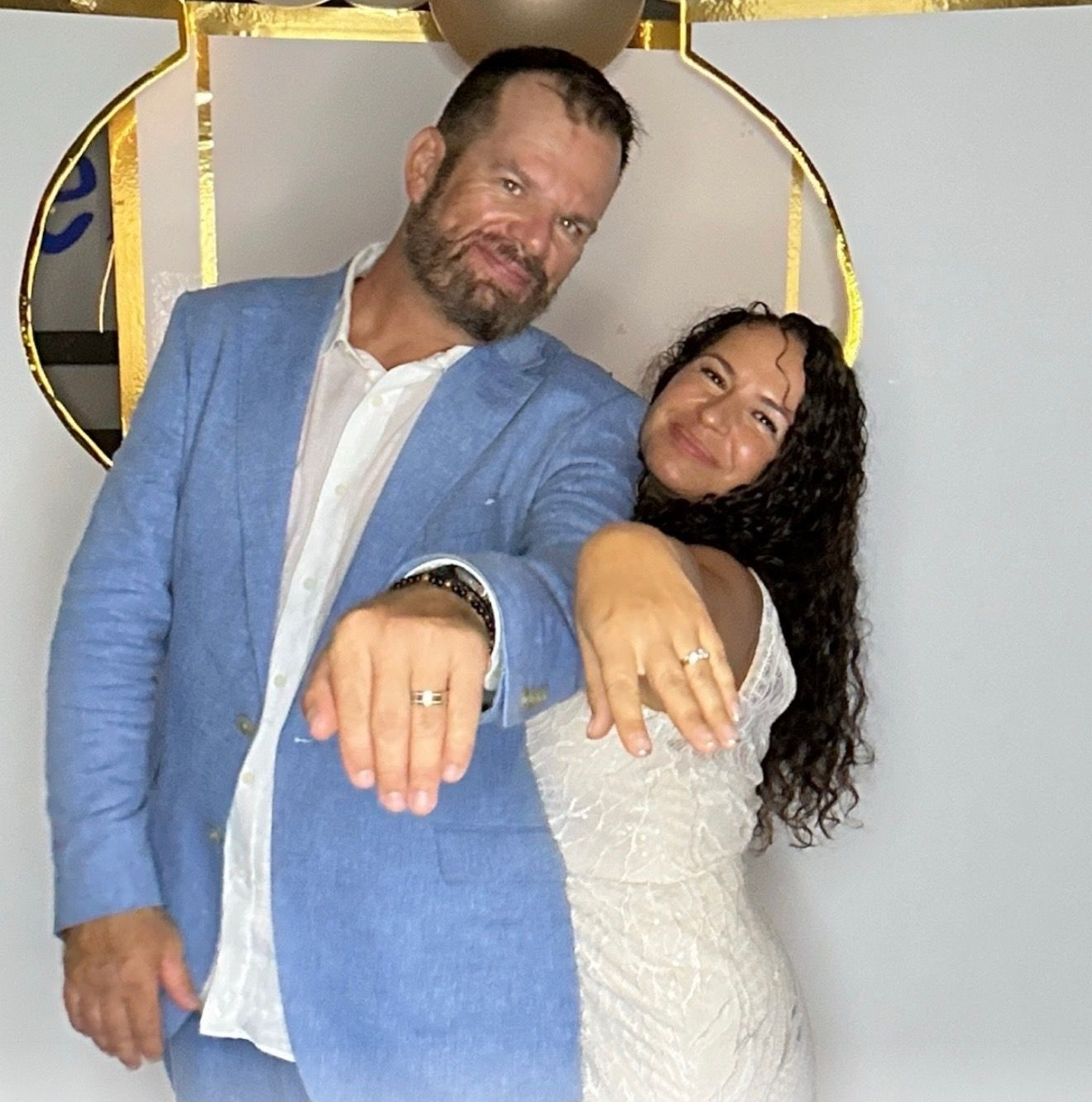 Couple smiles on a sunny beach; man has arm around woman, who displays diamond ring.