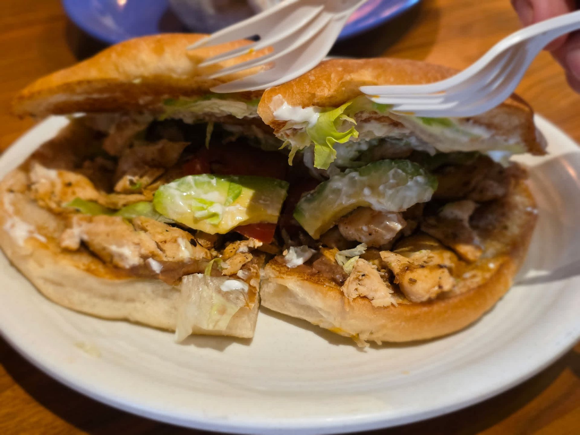 A sliced sandwich with meat and lettuce on a white plate, held open by plastic forks.