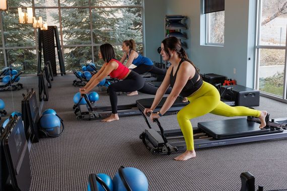People exercising on Pilates reformers in a bright studio, with medicine balls nearby.