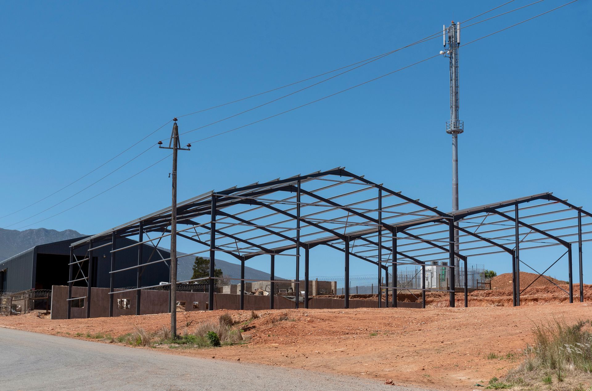 A partially constructed pole barn frame with metal posts and trusses, set against a clear blue sky on a sunny day.