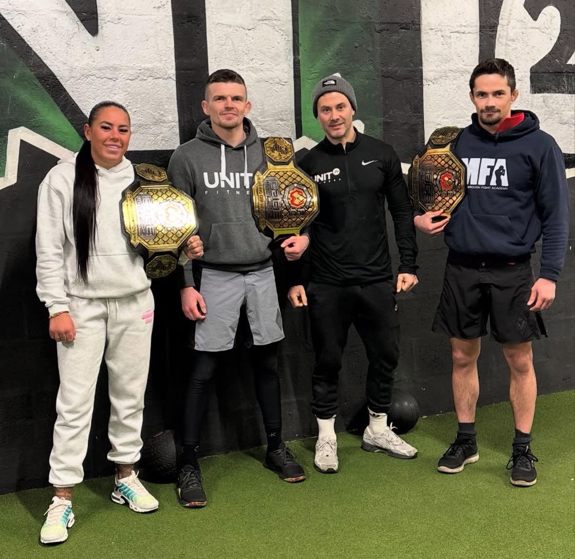 Four people pose with championship belts in a gym. Two wear gray sweatsuits. Two others in athletic wear.