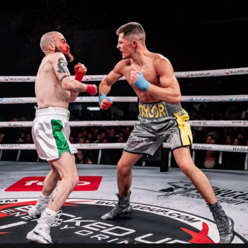 Boxers in ring: one throwing a punch at the other, who is bleeding. Both have gloves and shorts.