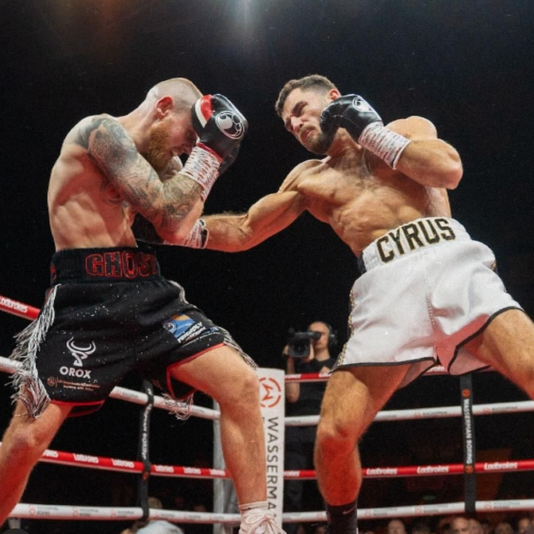Boxers in a ring, one throwing a punch. Black and white shorts, gloves up. Crowd in background.