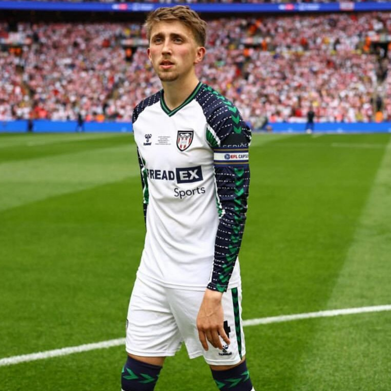 Soccer player in white uniform stands on field, looking towards the camera. Crowd visible in background.