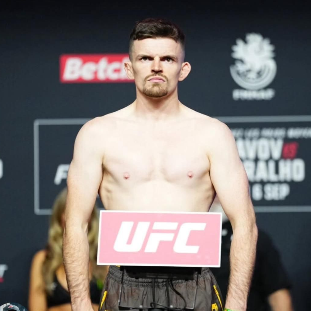 Man standing shirtless, holding a UFC sign, looking forward with a serious expression in front of a backdrop with logos.