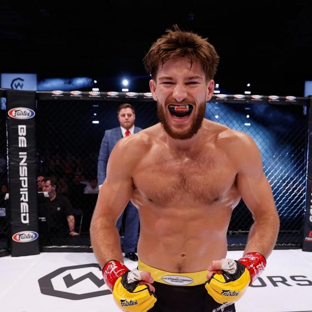 MMA fighter in ring, shirtless, mouthguard in, excited expression, gloved hands raised.