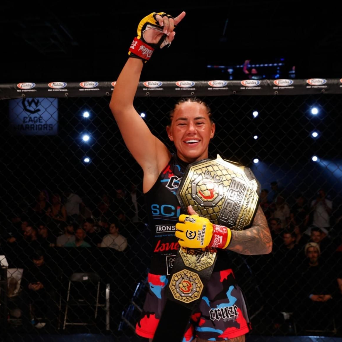 Female MMA fighter holding championship belt, raising arm, smiling in ring.