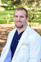 Man in a white lab coat smiles outside. Blue scrub top visible.