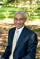 Man in a navy suit and gold tie smiles outdoors in front of greenery.