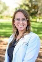 Woman in glasses and white coat smiles outdoors.