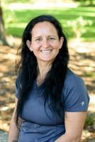 Woman with long black hair smiles, wearing a blue shirt, outside with blurred green background.