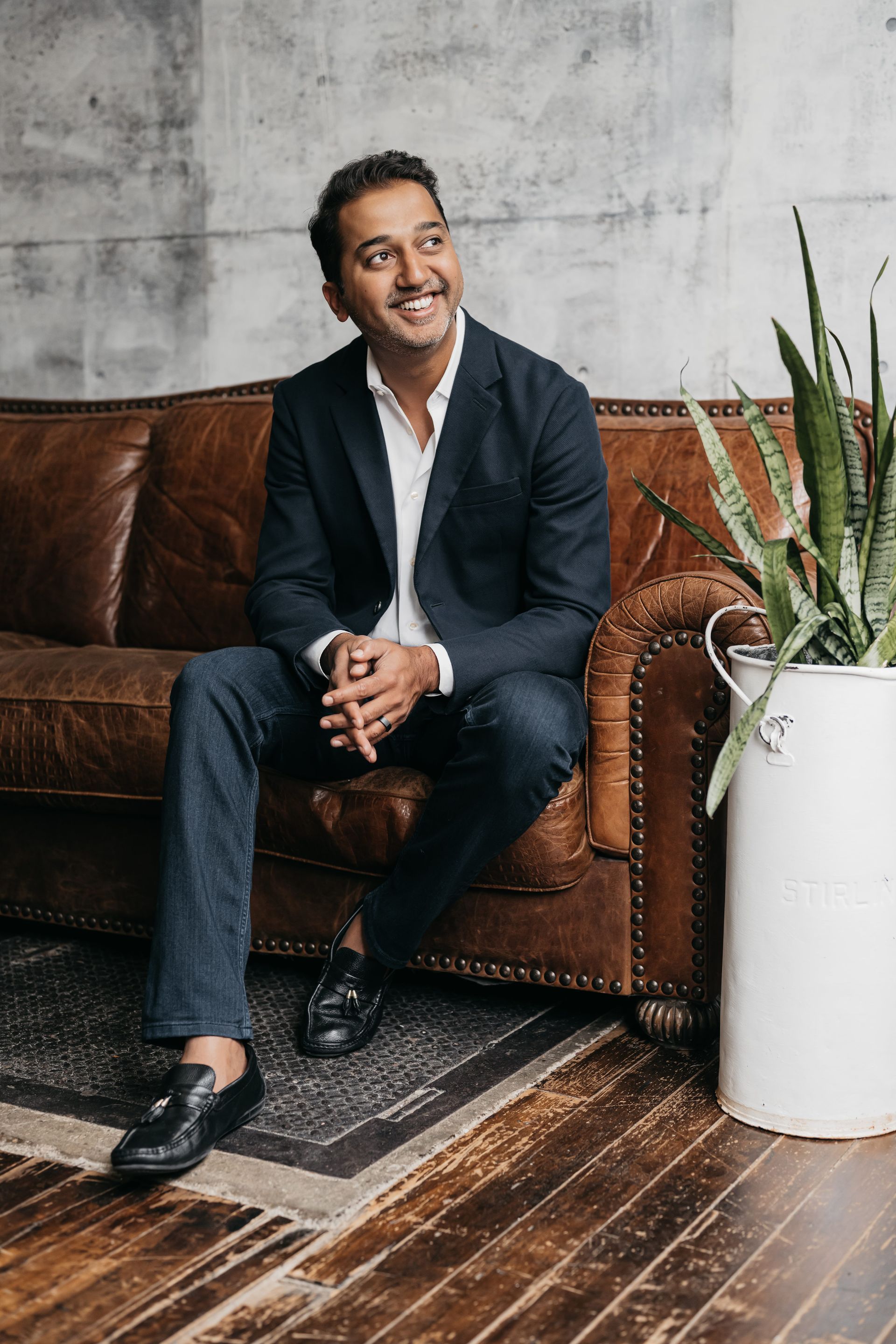 A man sits on a couch next to a potted plant, his left knee bent down, while his right leg stretches out. He has a small smile and a calm, confident demeanor.